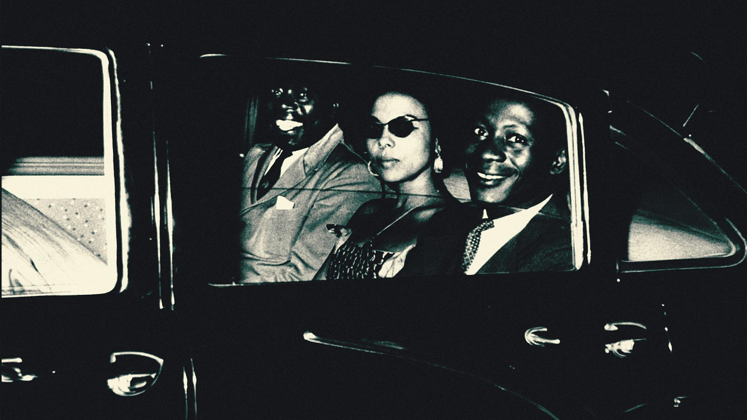 A black and white shot of a car featuring Andree Blouin (Patrice Lumumba's adviser) flanked by two men.