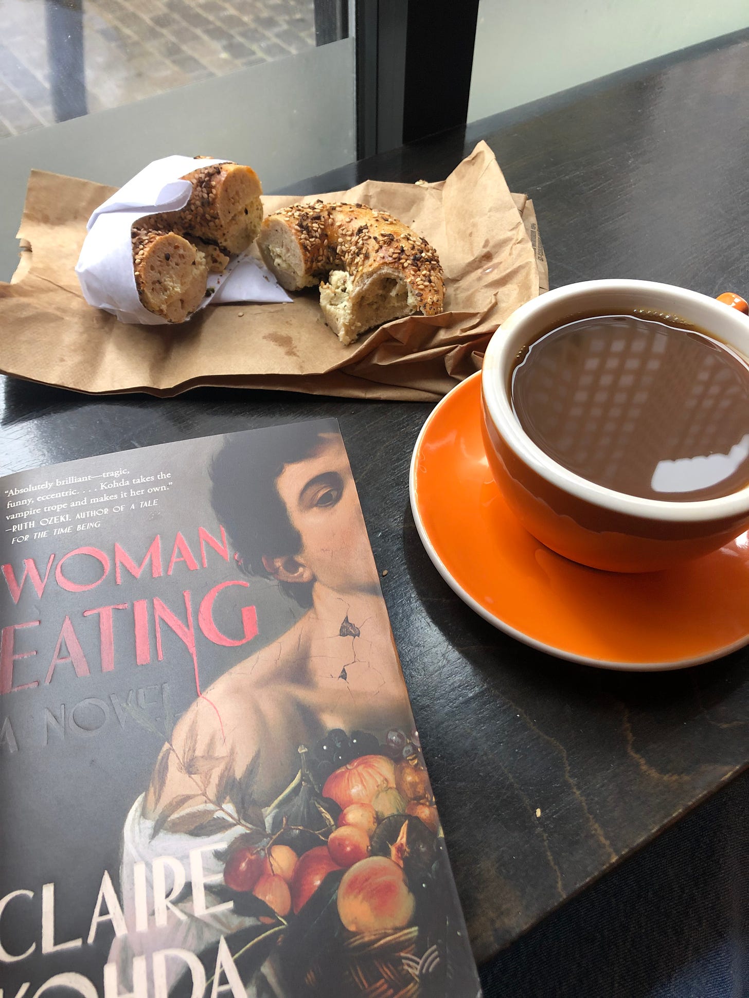 An image from a Boston cafe. An orange mug and saucer filled with coffee is on the right hand side of the image. In the top left corner there is a bagel and the bottom left sits a book called "Woman Eating"