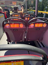 An open air, top deck of a bus. There are a few empty seats and then a man and child sitting towards the front of the bus. The area outside is fill of trees and the sun is shining.