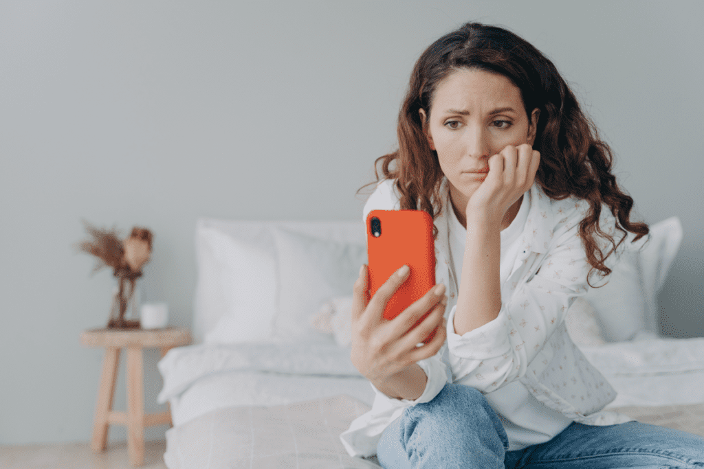 young-woman-sitting-on-edge-of-bed-focused-on-phone-screen-with-worried-emotions-and-stress-thinking-expression