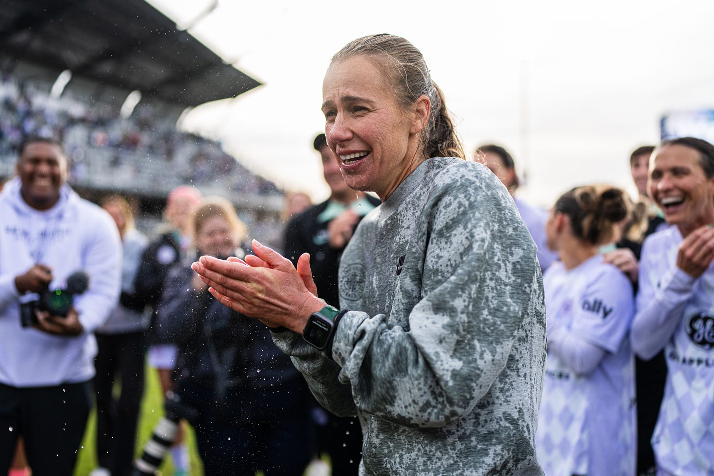 Women in wet sweatshirt smiles and claps while surrounded by smiling players and photographers