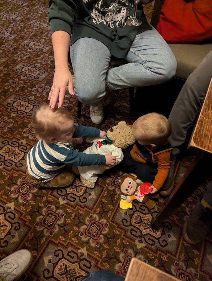 Two babies sat on a patterened pub carpet playing nicely with a teddybear. One of their mums is paying close attention.