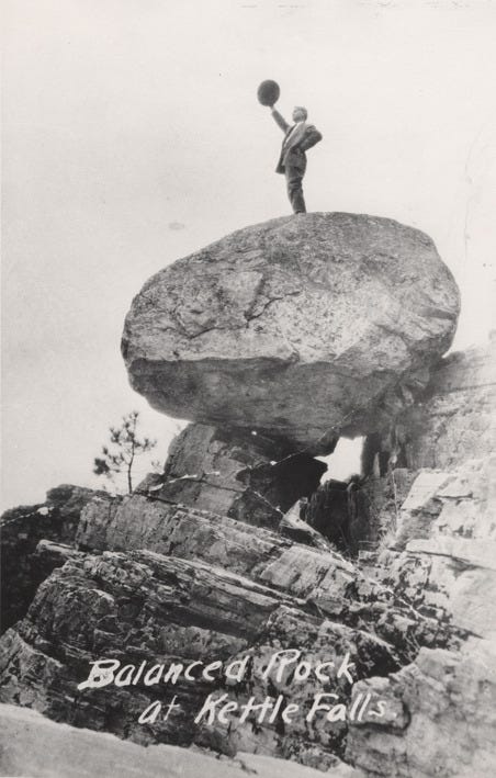 Black and white photograph of a man standing on top of a rock balanced on top of other rocks