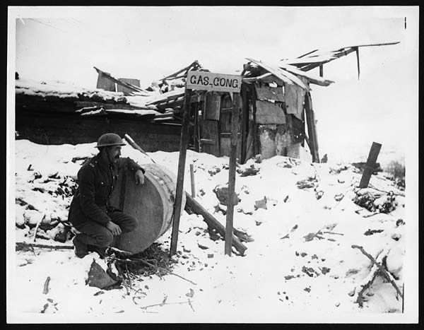 Black and white photo of a British soldier sat next to an oil drum underneath a sign saying "gas gong". He's in the snow and looks watchful but a bit bored.
