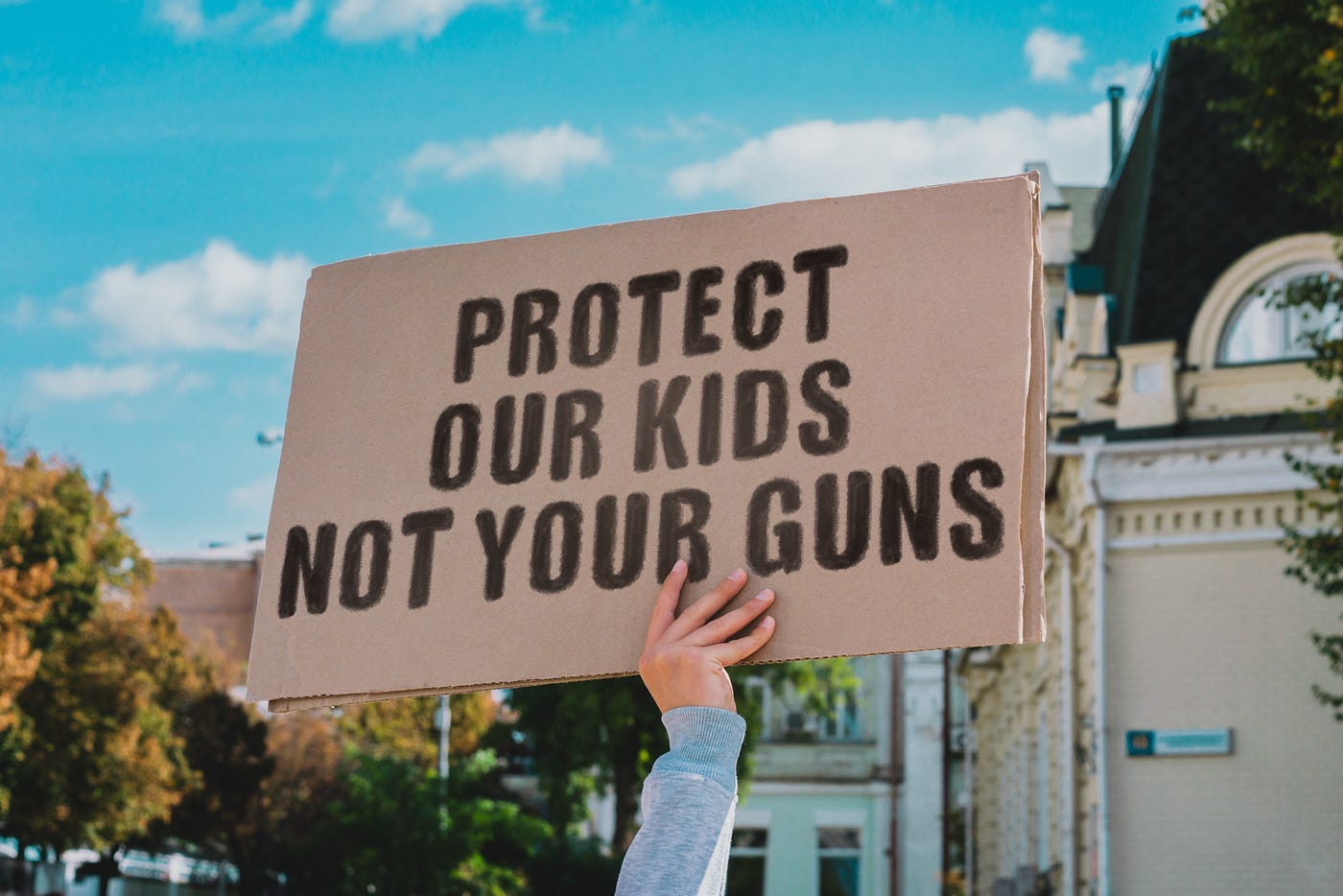 The phrase “ Protect our kids not your guns “ on a carton banner in men’s hand. Human holds a cardboard with an inscription. Gun violence. Law. USA. Ban
