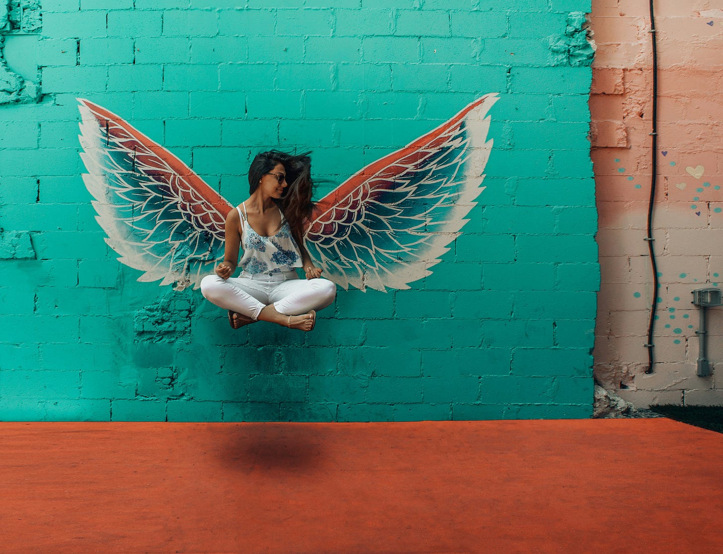 Woman with brown skin and long black hair wearing skinny white jeans and spaghetti-strap white-and-blue floral top sits cross legged on a hidden ledge with colourful angel wings painted on the wall behind her. Wall is turquoise and ground is rust-brown. She's wearing sunglasses and looking to the right.