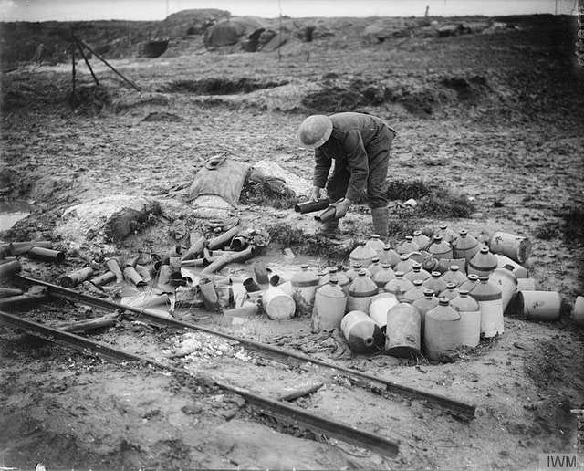 A black and white photo of a pile of rum jars and shell cases. A soldier is holding two shell cases as if he’s sorting them out.