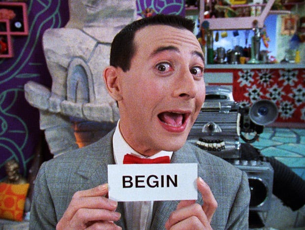 Picture of a white man in a suit on a TV set wearing a red bow tie and a grey suit. He is holding a card that shows the word "BEGIN."
