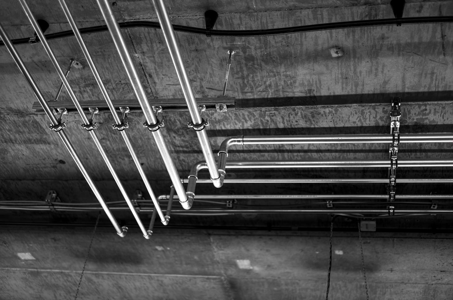 Black-and-white photo of five metal water pipes running along a ceiling. The pipes each turn and curve in an elbow, changing from running left/right to running up/down