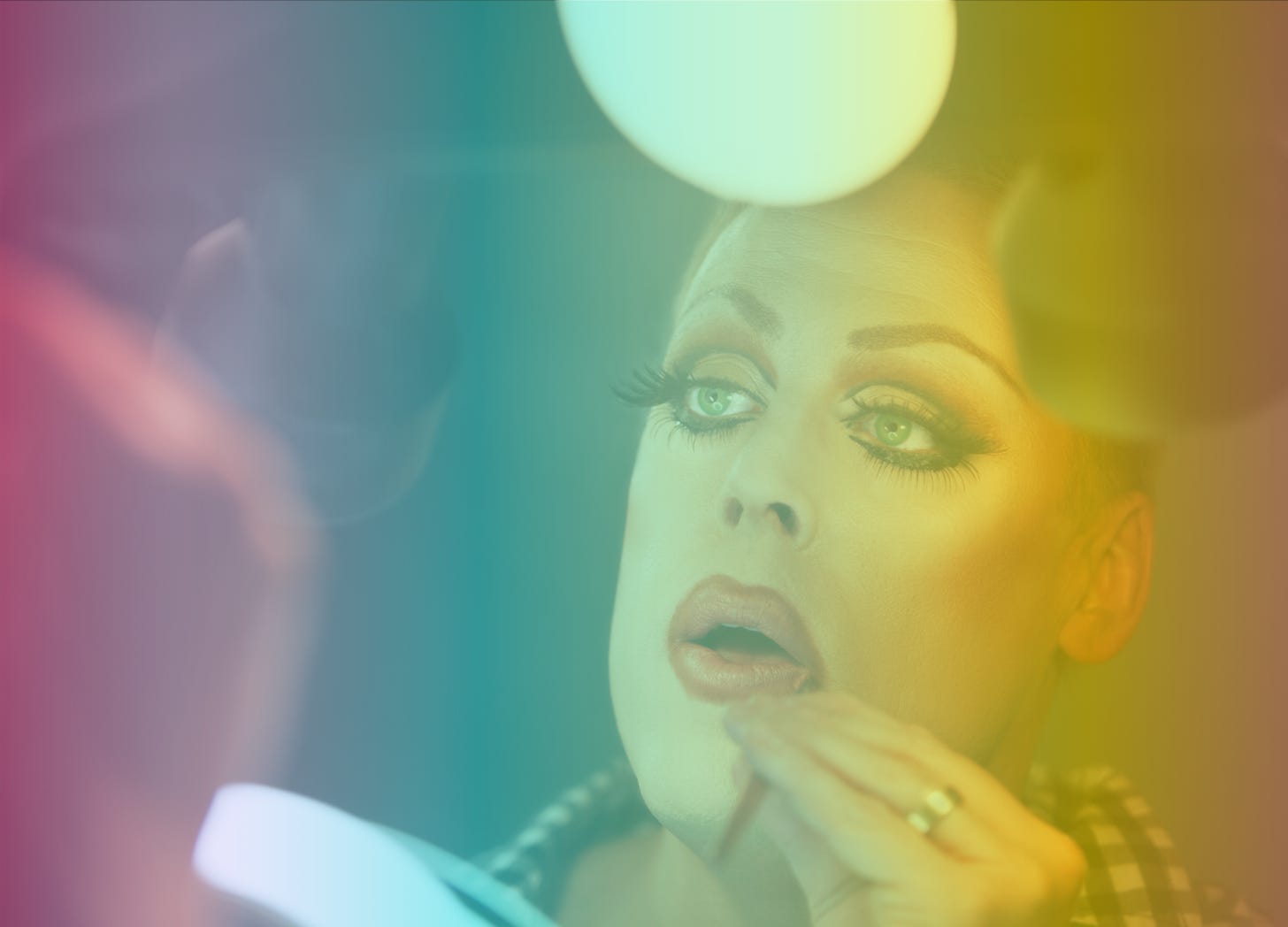 A rainbow-filtered image of a drag queen putting on makeup in the mirror