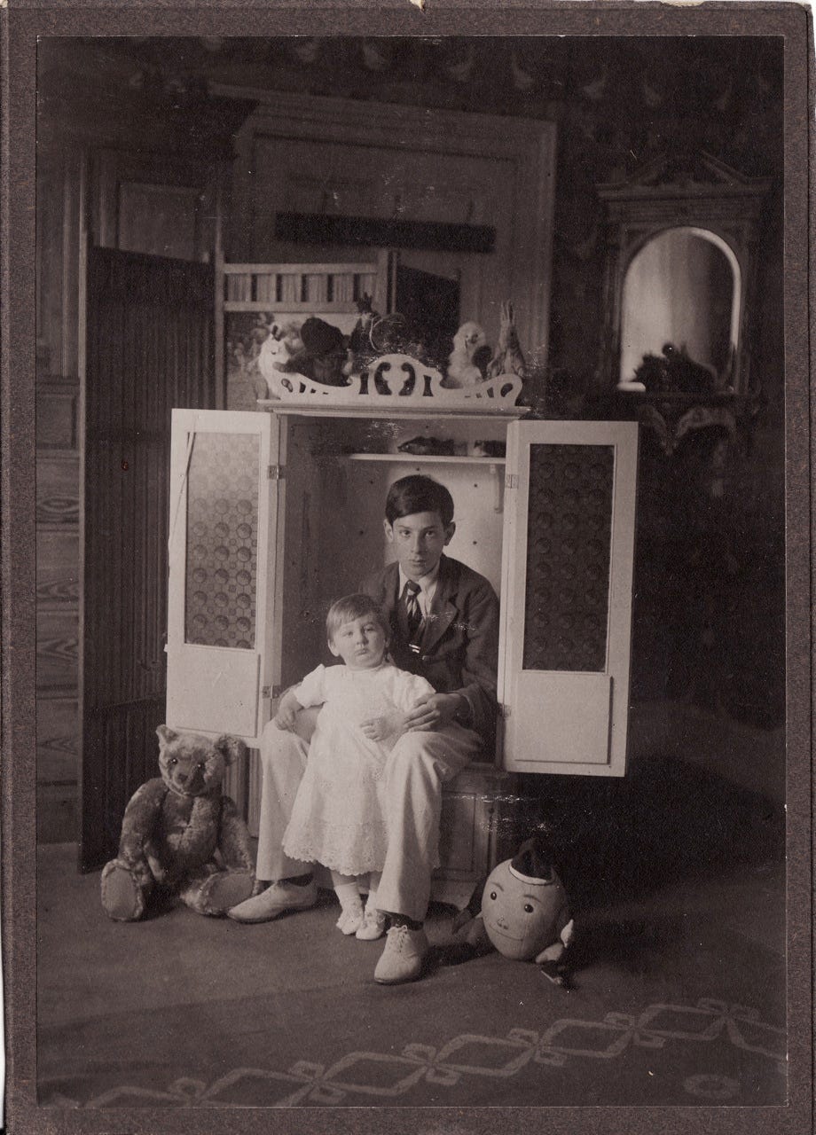 A vintage photograph from around 1930; it's black and white and depicts two children posing inside a cupboard. It's a formal portrait and there are teddy bears and toys arranged around them.