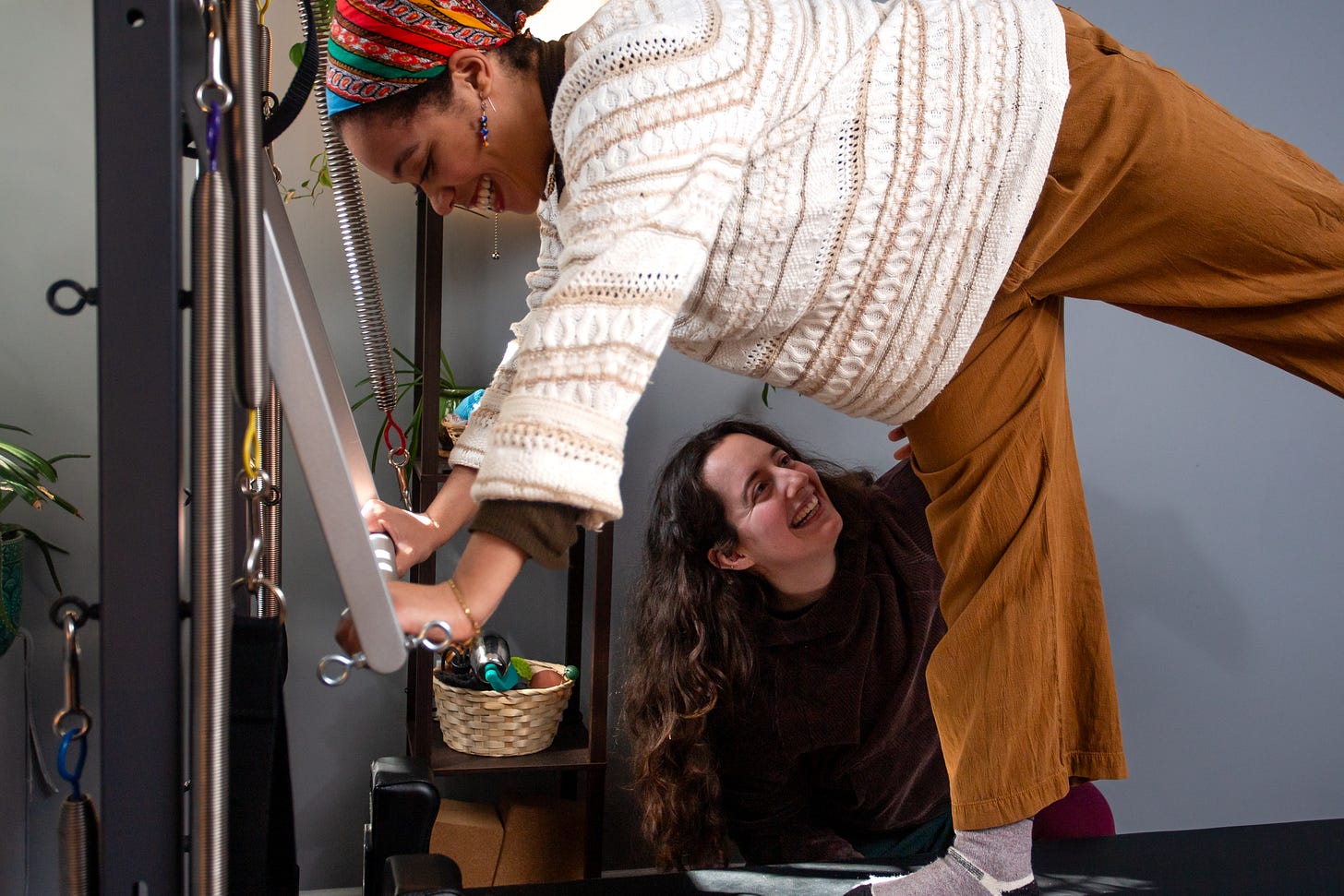 Sofia guides a client through movement on the Pilates reformer, placing a hand on the client's thigh. They are both smiling.