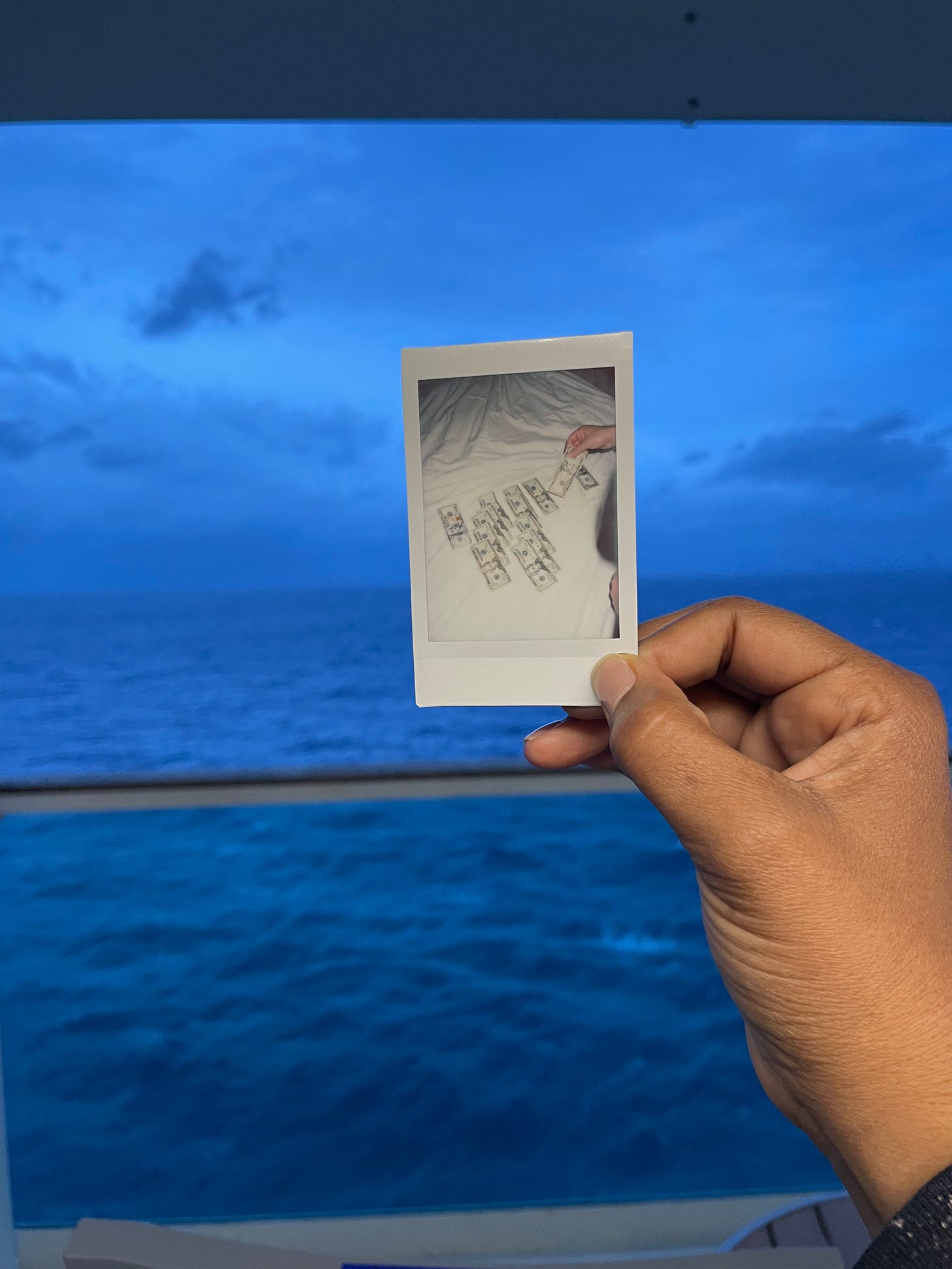 a mini polaroid of a money spread out on a bed, held up against the background of the ocean view from a cruise ship balcony. 