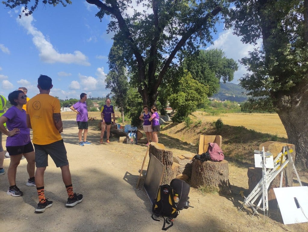 After the event, some purple-wearing participants chat, while the event paraphernalia (a message board and some poles) rest on some tree stumps. To the right of the path are a few trees, mostly hiding the view of the hills behind.