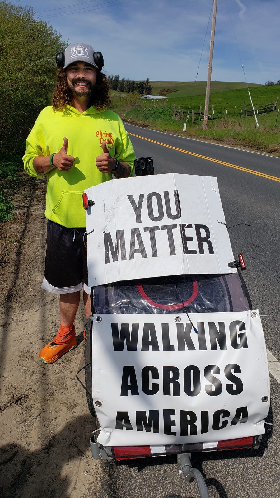 A young white man with curly shoulder-length hair wearing a ball cap, headphones, a green flourscent shirt, pushing a cart with 2 signs - one says You Matter, the other says Walking Across America