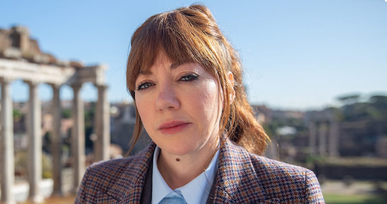 Photo of fictional TV host, Philomena Cunk. In the background are some archaeological ruins