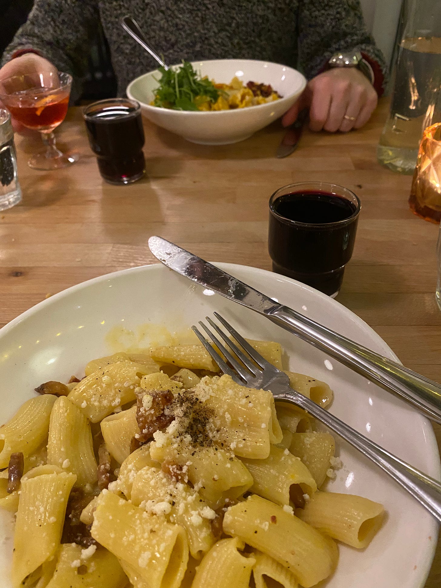 In the foreground, a shallow white bowl of rigatoni carbonara with parmesan on top. There's a glass of red wine on the table above and to the right, and a fork and knife on the edge of the bowl. Across the table in the background, Jeff has his hand on a glass of a half-empty cocktail, and a fork is sticking out of his bowl of pasta, which is covered in arugula.