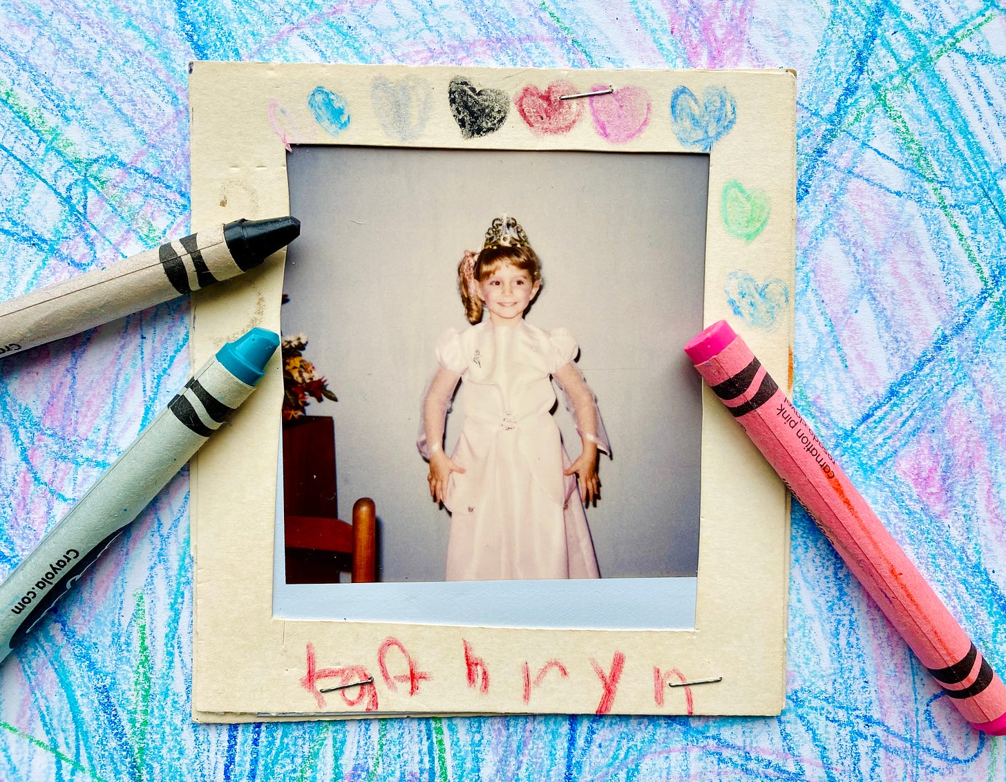 a polaroid photograph of a 4-year-old child in a princess costume. the child is smiling but looks uncomfortable. the photograph is surrounded by a paper frame, on which the child has drawn hearts in crayon and scrawled “kathryn.” the photo rests on a piece of paper covered with crayon marks, and three crayons are arranged around the photo’s edge.