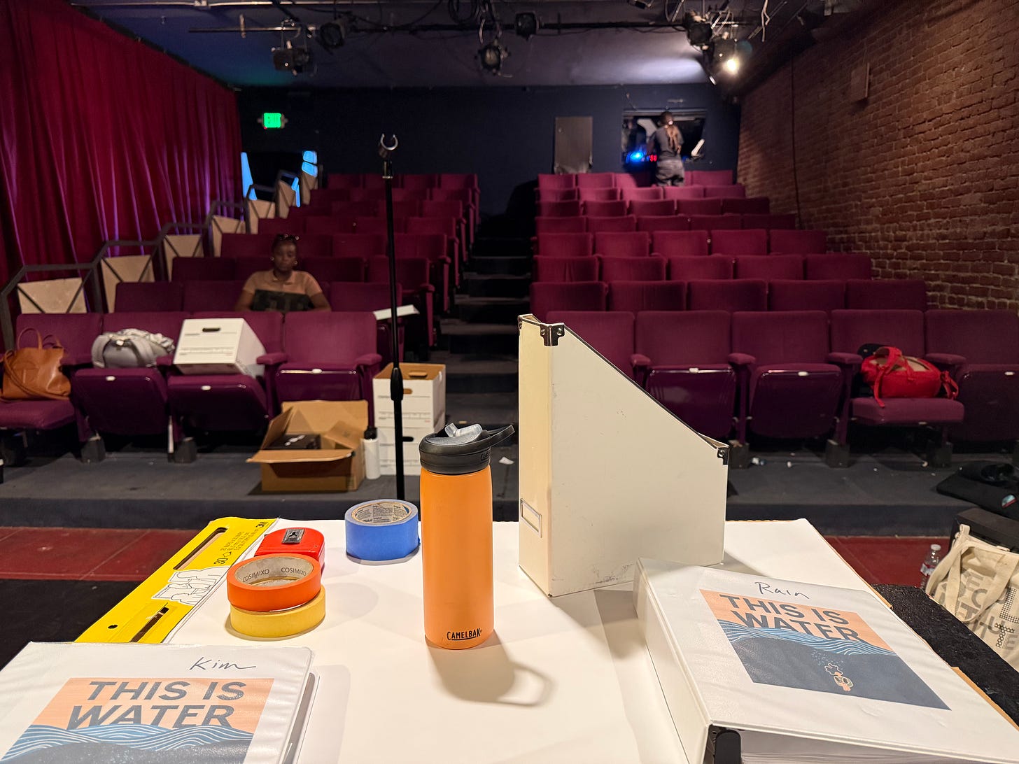 the view from the stage of a small theater with a table in the foreground covered in scripts, painter's tape, water bottle - and in the seats a young Black woman on her computer the view from the stage of a small theater with a table in the foreground covered in scripts, painter's tape, water bottle - and in the seats a young Black woman on her computer