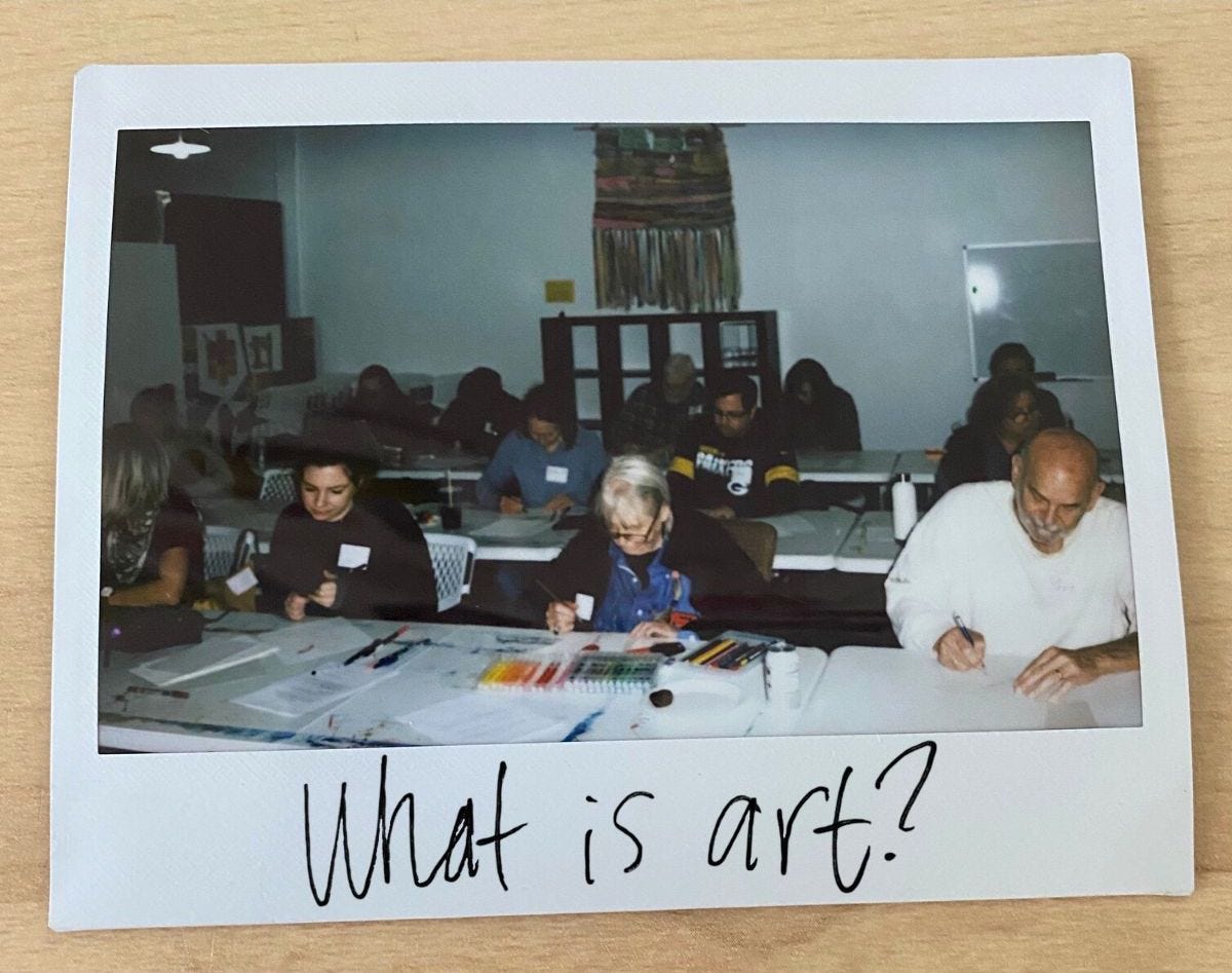 A polaroid picture of the class, with 4 women working on the front row, a man and 3 women on the second row, and a man and woman working on the back row. Rainbow markers and white paper are sitting on the table in the front row. On the bottom of the polaroid is written "What is art?" in black pen handwriting.