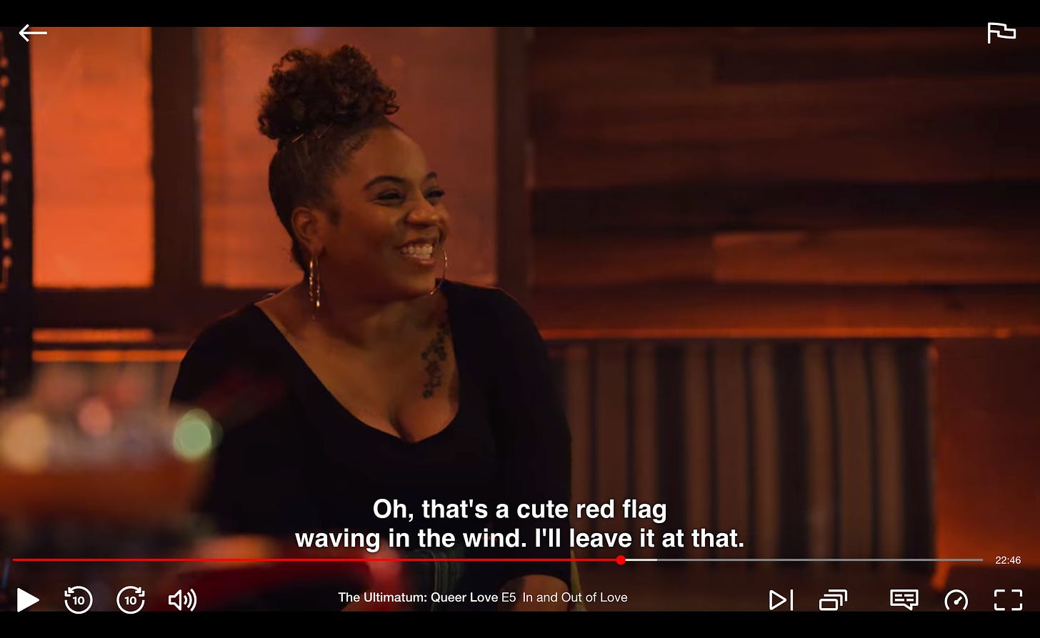 A screenshot of the Netflix reality show The Ultimatum: Queer Love. A Black woman is sitting in a bar and smiling with gritted teeth. Below her is her dialogue: "Oh, that's a cute red flag waving in the wind. I'll leave it at that."