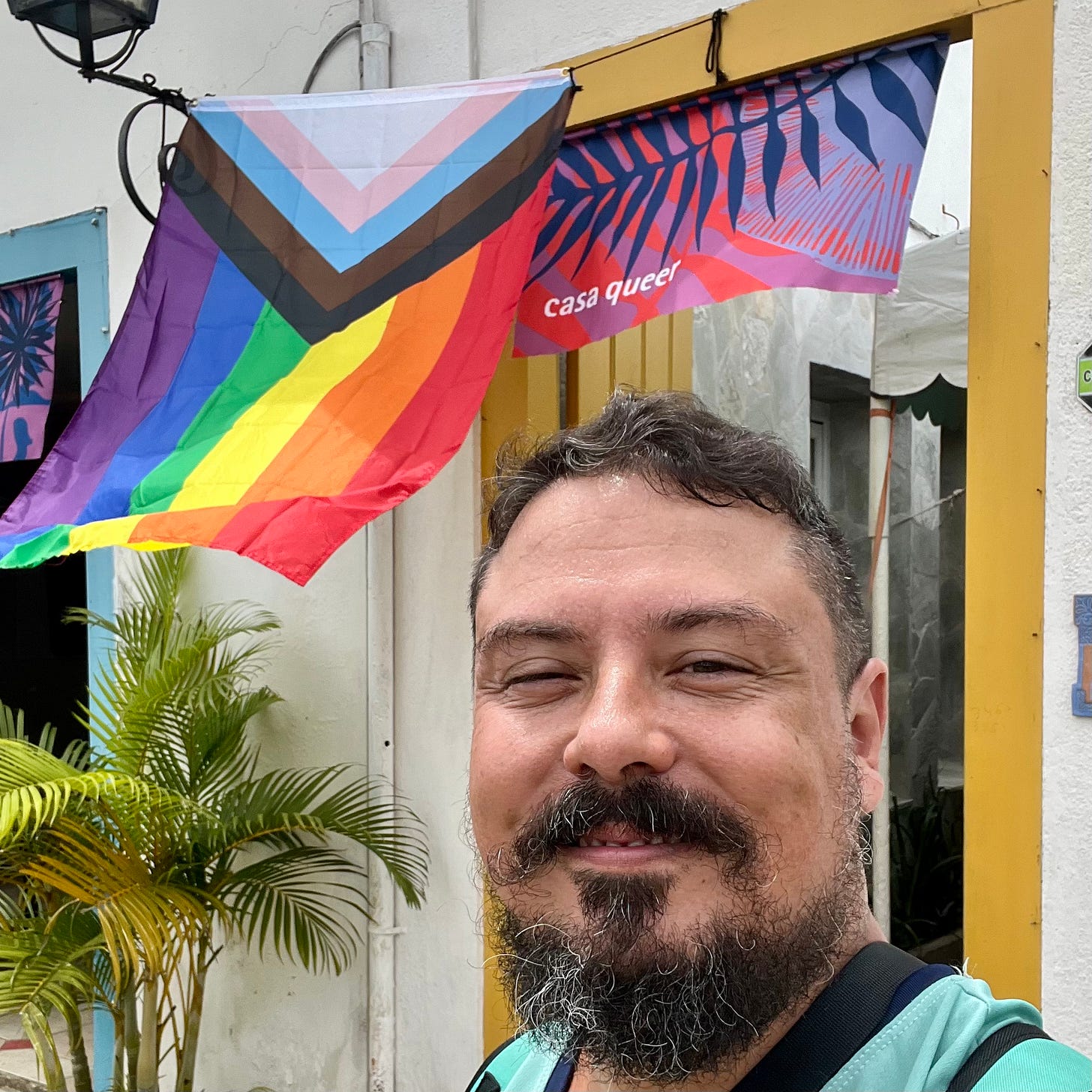 selfie minha em frente à casa queer. sobre a porta há uma bandeira progressista do orgulho e uma bandeira com o nome da casa. Sou um homem de pele bege-clara, olhos, cabelos, barba e bigode castanhos. Tenho fios brancos na barba e sou gordo.