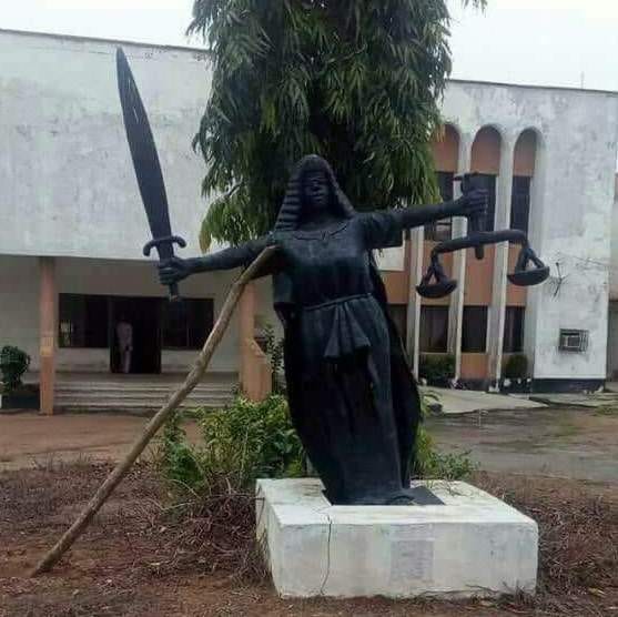 The image shows a status of justice in Nigeria, leaning to the left and being propped up with a stick