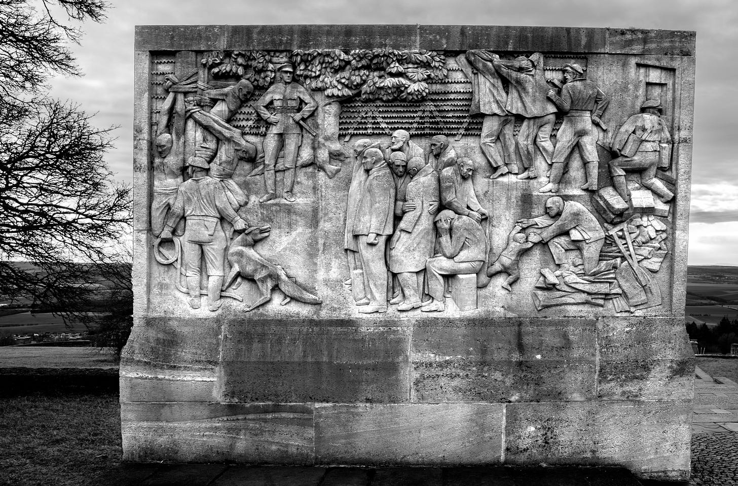 A black-and-white photograph of the Buchenwald Memorial of the GDR, a relief sculpture depicting scenes of oppression and suffering within the Buchenwald concentration camp. The carved figures show a group of individuals enduring harsh treatment: on the left, soldiers oversee the scene with weapons and dogs, while people are bound or forced against a wall. In the center and right, grieving civilians are depicted, including women holding children and people in despair. The artwork reflects the harsh realities of camp life but carries the theme of resurrection, symbolizing the enduring human spirit amid profound injustice. The background features symbolic patterns and trees, adding depth to the somber narrative of oppression and resilience.