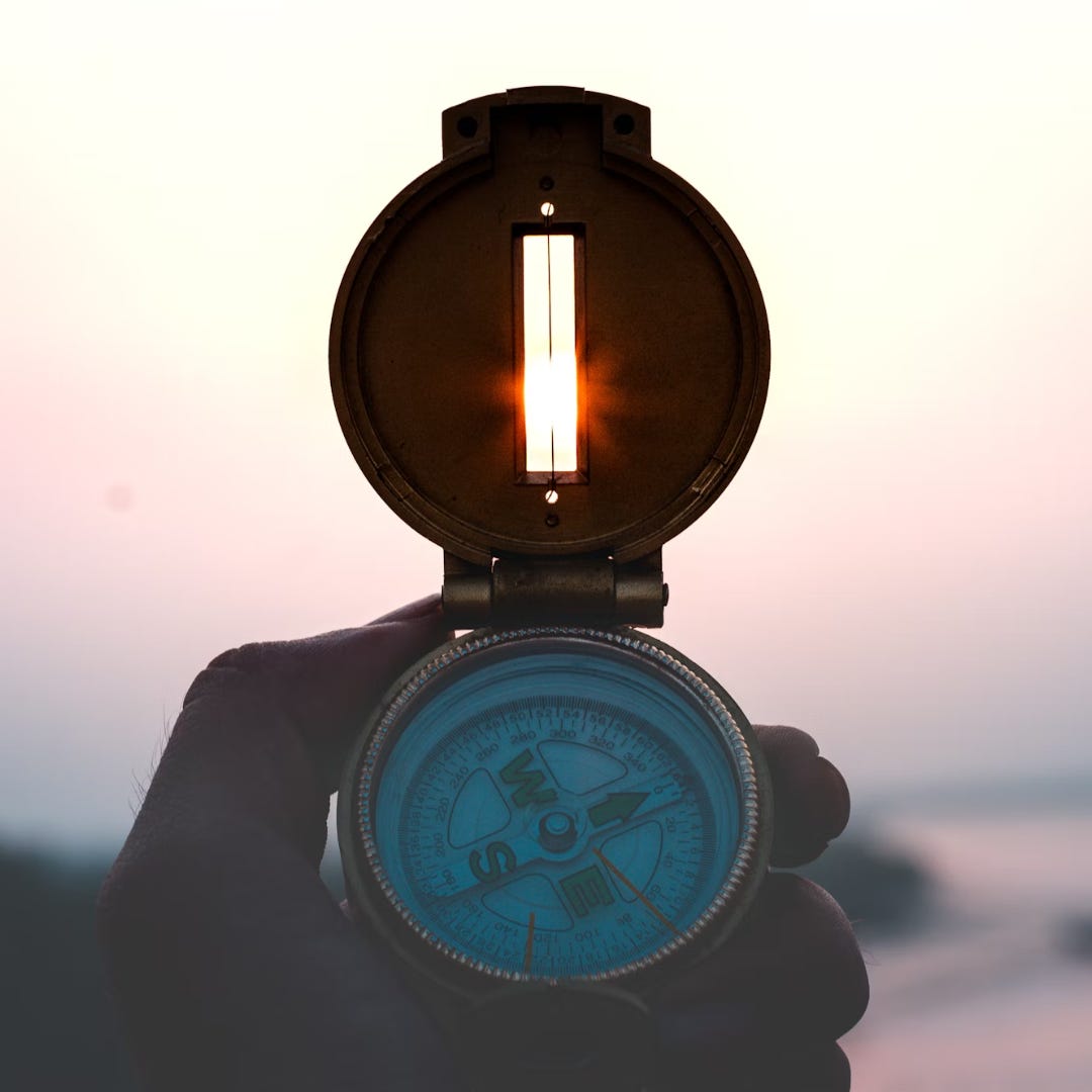 A close-up of a compass held-up to the horizon line. The sun low on the horizon shines through an open slot in the compass’s cover.