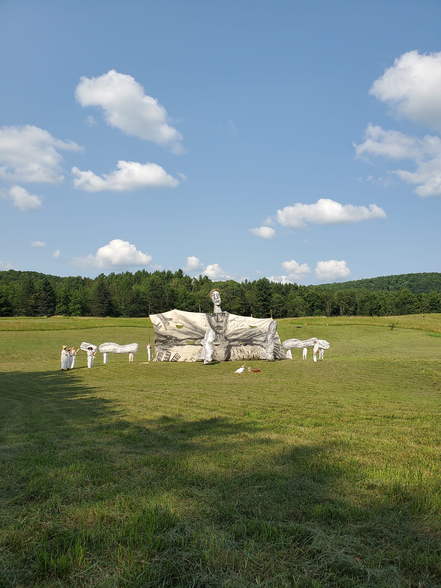 A humanoid figure is assembled from limbs and hands against a massive banner, all held aloft by human puppeteers, dwarfed by the art and by the landscape--blue sky and clouds, an open green field, and woods looming beyond at the horizon.