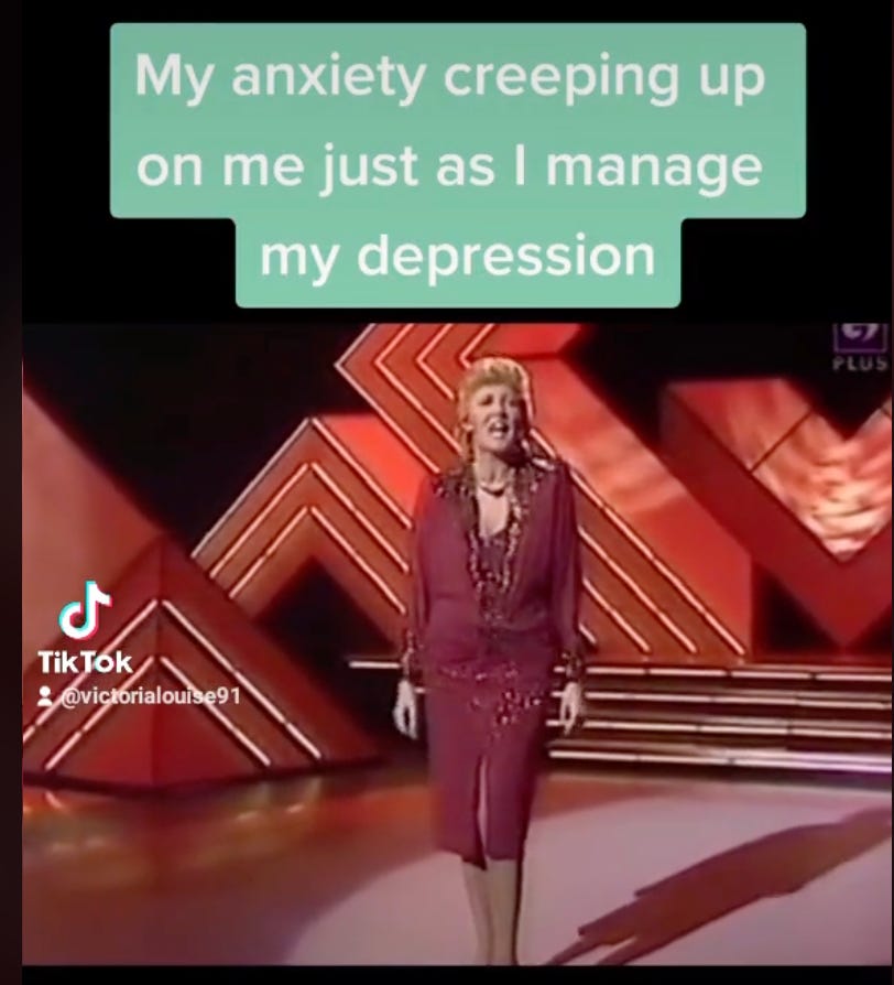 A screenshot of a TikTok video featuring a woman standing on a stage with a retro design in the background. She is dressed in a vintage purple outfit, singing with an expressive face. Overlaid on the video is a caption in a speech bubble that reads 'My anxiety creeping up on me just as I manage my depression'. 