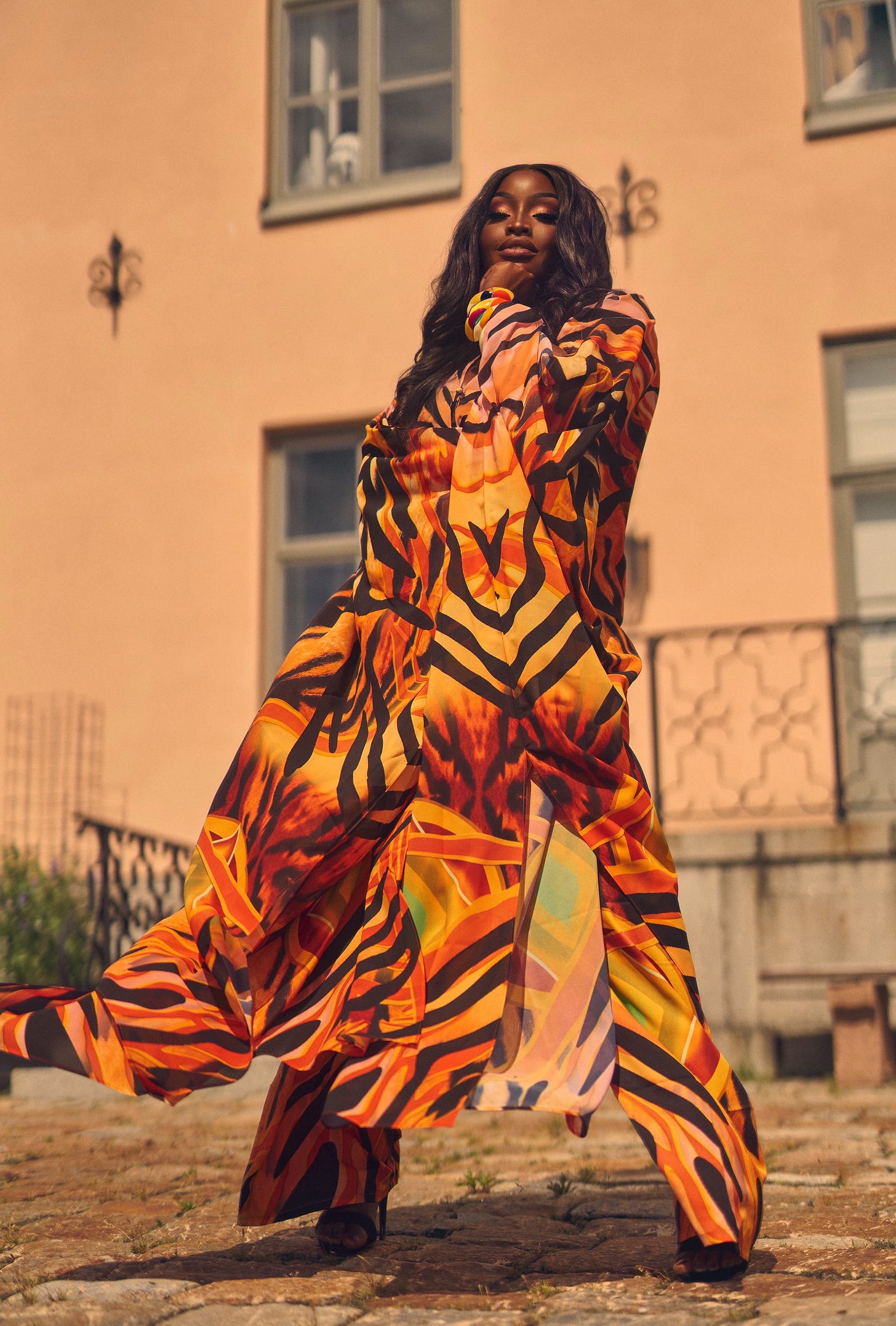 Lovette Jallow posing powerfully in a vibrant orange, black, and yellow patterned outfit, standing against a sunlit peach building, captured during a fashion-forward outdoor shoot in Sweden.