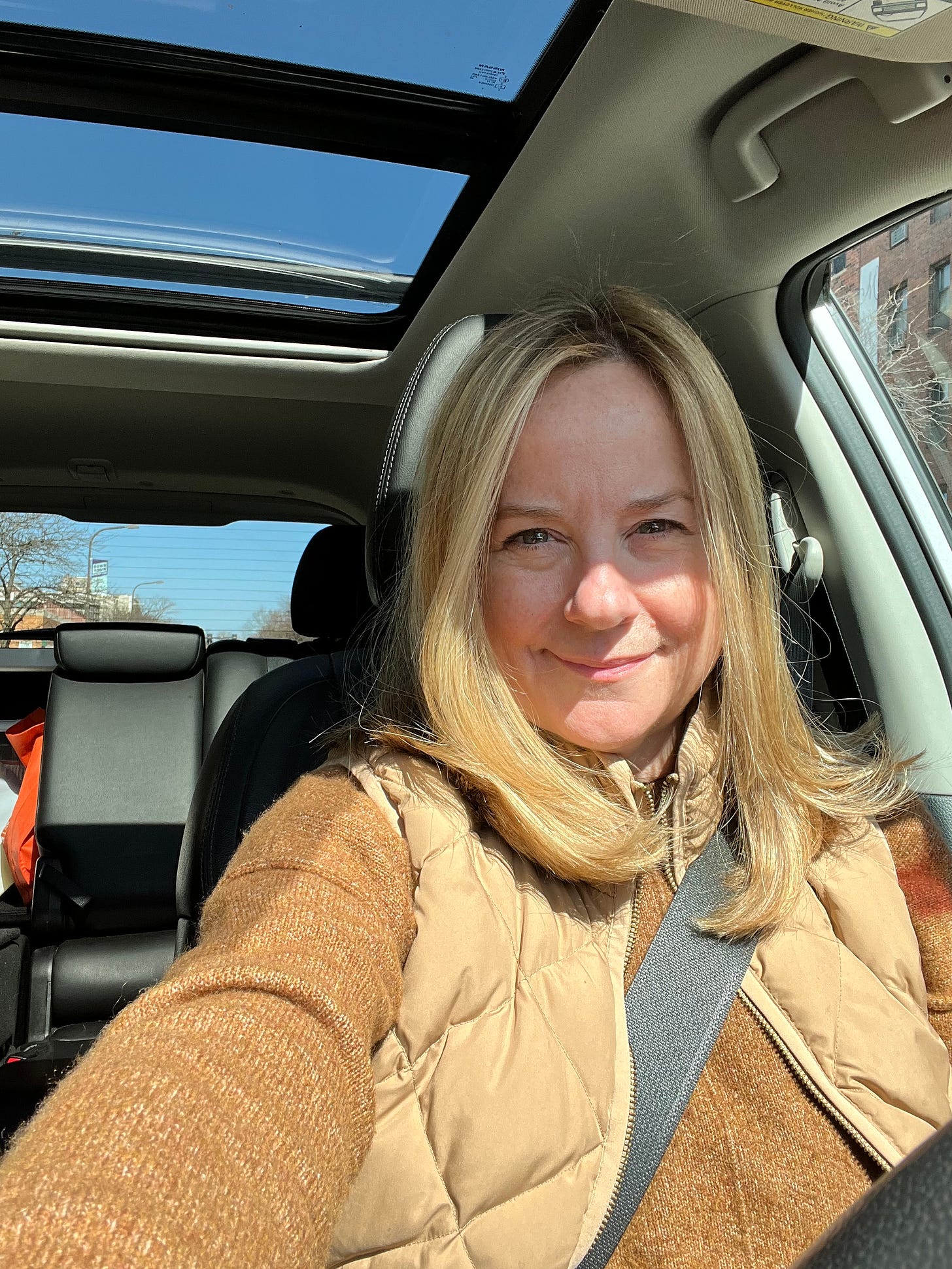 Blonde woman in a car with a sunroof. Blue sky can be seen through the sunroof. She's wearing a brown sweater and a lighter brown vest. 