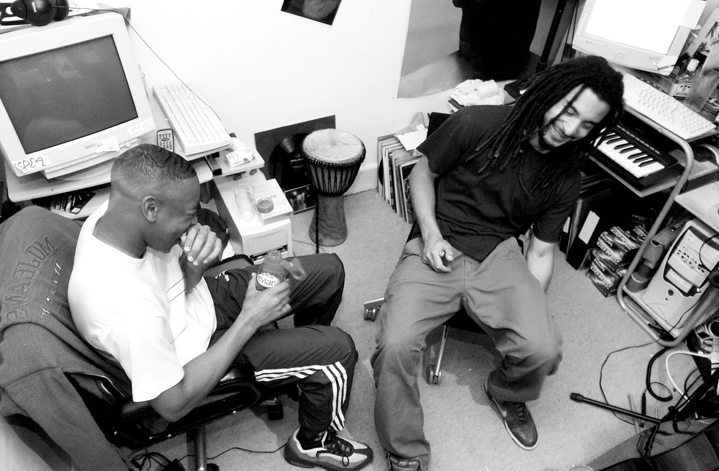 Two young men (Coki and Mala) sat on office chairs laughing in a home music studio surrounded by computers, instruments and other related objects