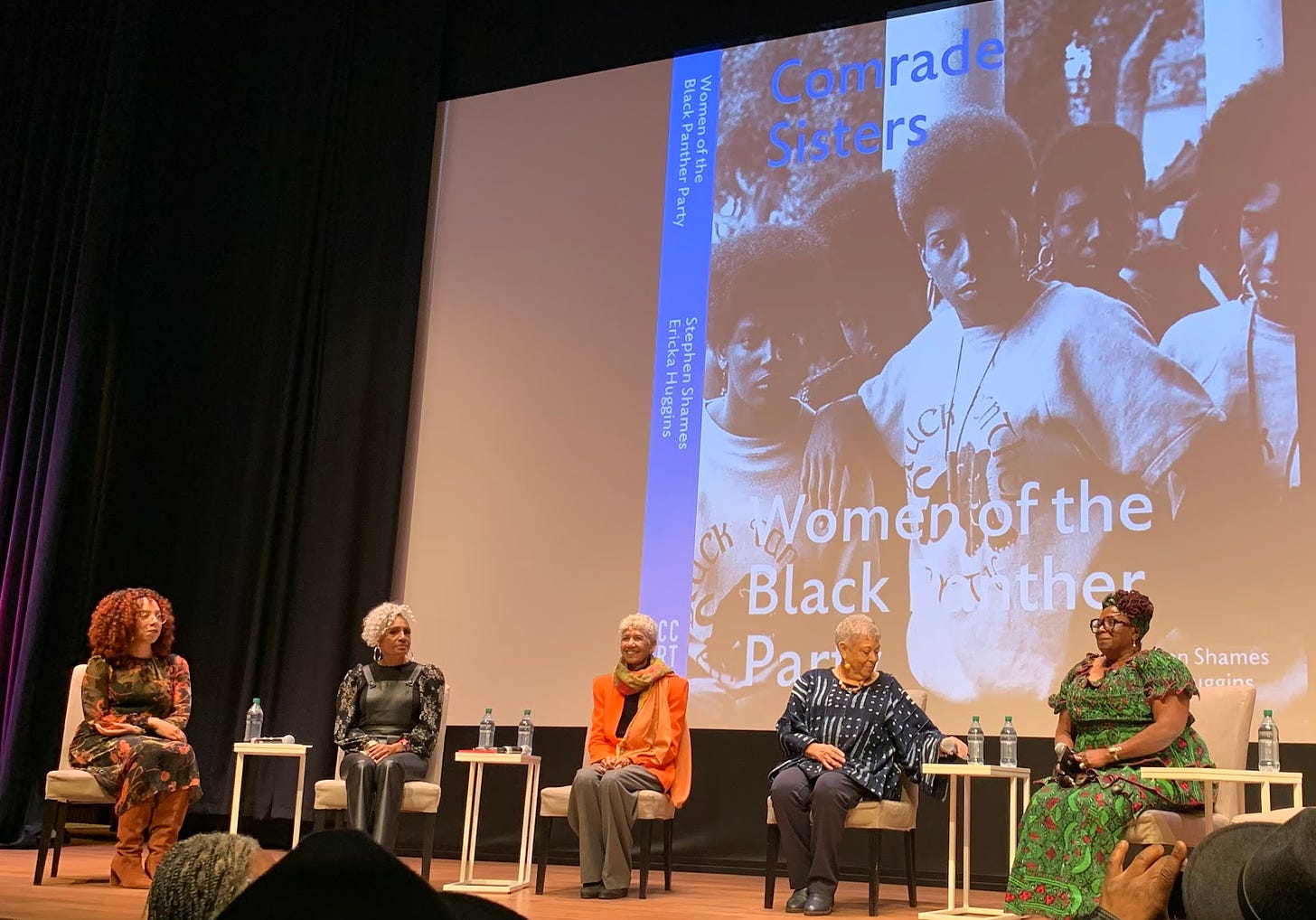 Five Black women seated on a stage. Behind them is a large screen with a black and white photo of Black people with afros wearing Black Panther t-shirts. Words on the screen read, “Comrade Sisters” and “Women of the Black Panther Party.”
