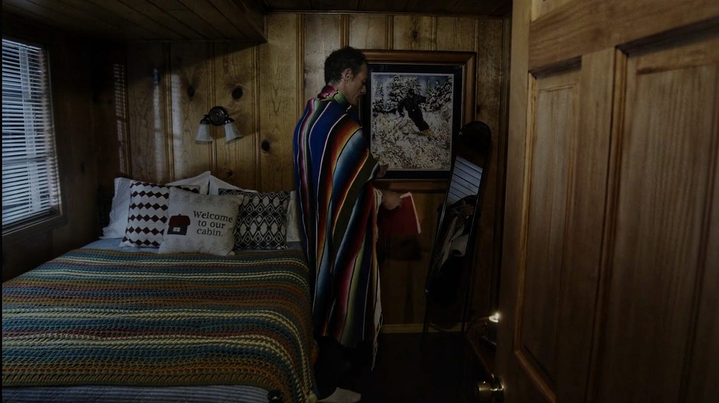 Desmond, wrapped in a striped blanket, in his wood-paneled bedroom. There is another striped blanket on the bed as well as a bunch of throw pillows, including one reading "Welcome to our cabin."