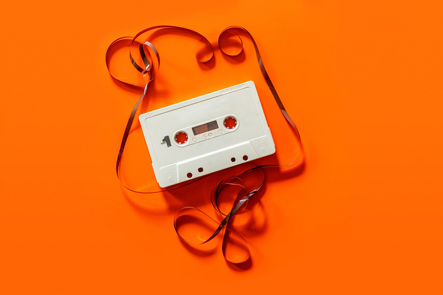 Su uno sfondo arancione, un'audiocassetta bianca con la bobina sfilata e annodata fuori dalla custodia. Su uno sfondo arancione, un'audiocassetta bianca con la bobina sfilata e annodata fuori dalla custodia.
