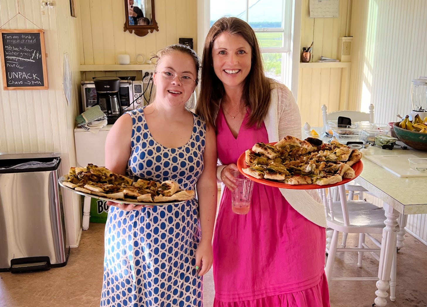 Penny and Amy Julia stand in a kitchen holding platters of food and smiling at the camera.