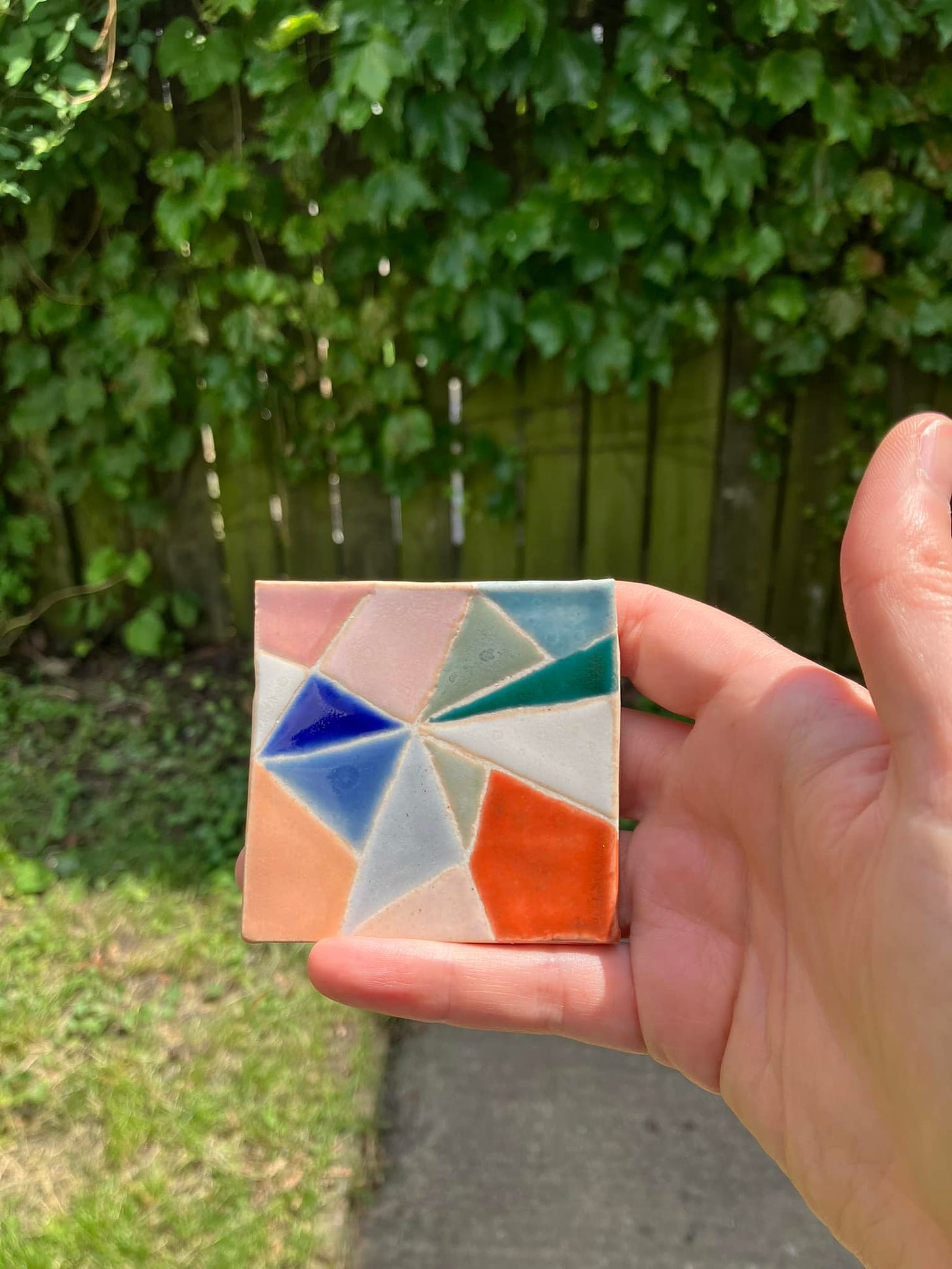 multicolored test tile with geometric design, held up in natural light, in front of an ivy covered fence