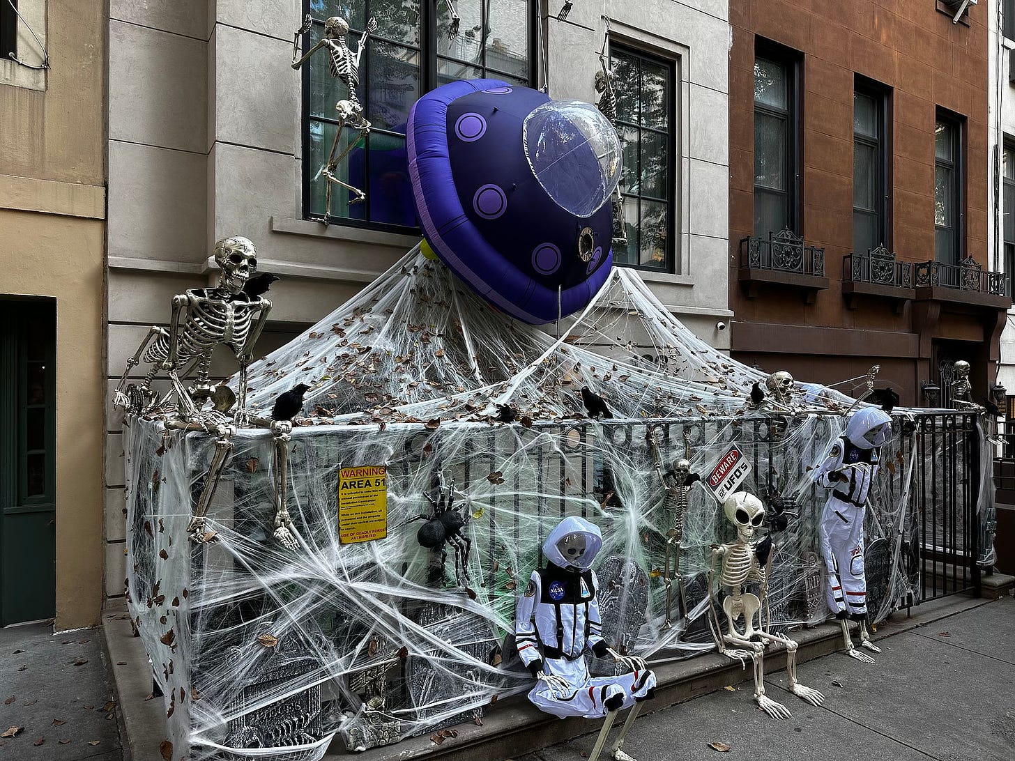 Photograph by Michael Whelan featuring the base of a build in NYC with metal fence wrapped in spider webs. Two skeletons in space suits sit next to a alien skeleton. A sign reads "BEWARE UFOS" while another says "WARNING: AREA 51". Silver skeletons climb over the railing and up the building as a inflatable UFO, blue and purple, crashes down.