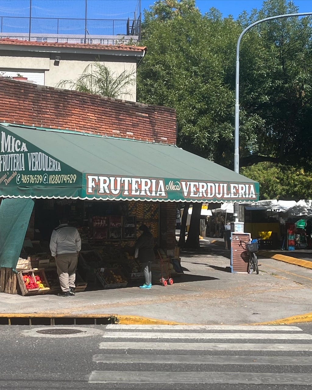 Fachada de uma banca de frutas e verduras numa rua de Buenos Aires. O espaço tem um toldo verde com a borda ondulada, em estilo antigo, com as palavras FRUTERIA VERDULERIA pintadas a frente