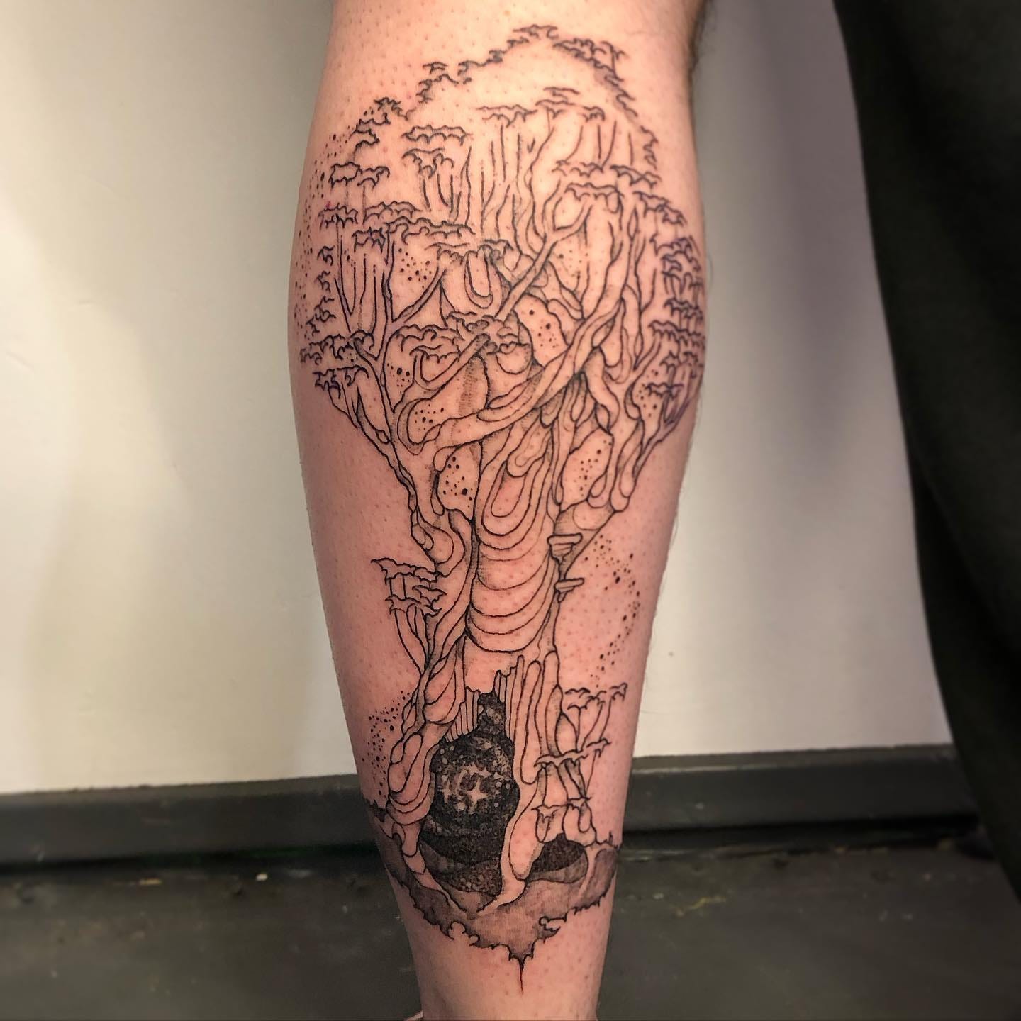 a tattoo for Ashley by bug on the back of her calf of a big tree with a sparkly void portal at the base she has a light skin tone and its done in black ink