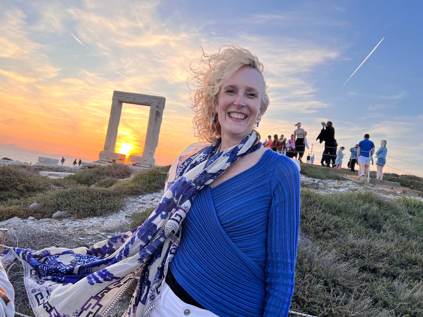 Rev. Jen Butler wearing a blue sweater and long scarf standing on a mountain with her hair blowing in the wind in front of a large square stone arch with a sunset in the background. 