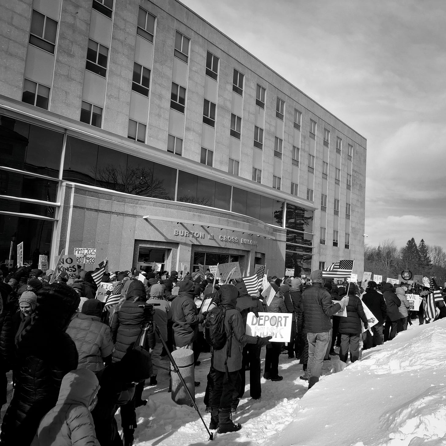 A crowd of protestors march through snow holding signs in front of the Burton Cross Building.