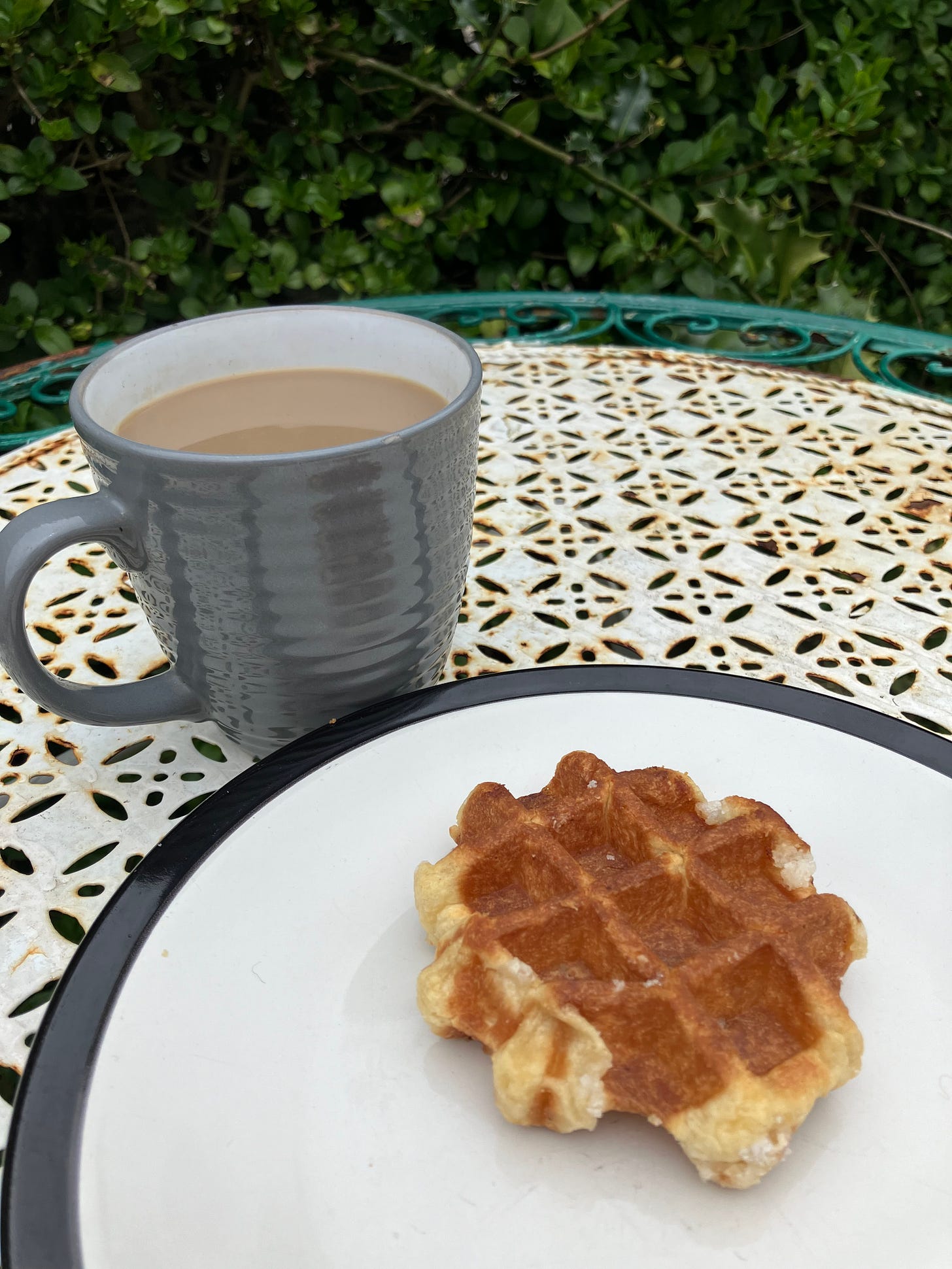 On a white and green garden table, sits a grey mug of coffee and a sugar waffle on a black rimmed plate.