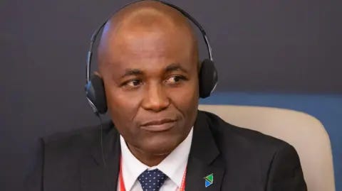 WHO African Region/X Faustine Ndugulile in a black suit and headphones and a Tanzanian flag lapel