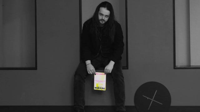 A man in black sits in a niche looking gloomily at the camera. In his hands is the zine, bright yellow in an otherwise black and white image.