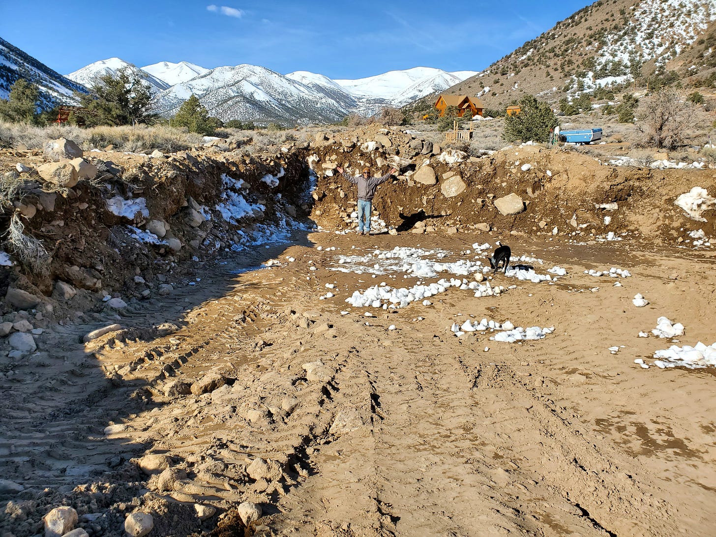 A view of a large excavated hole in the ground. Eric is standing in the far side of the hole with his arms outspread.
