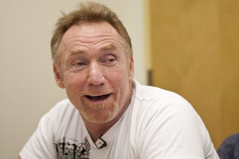 A man in a white t-shirt with red hair is facing the camera, looking to the right, and smiling. 