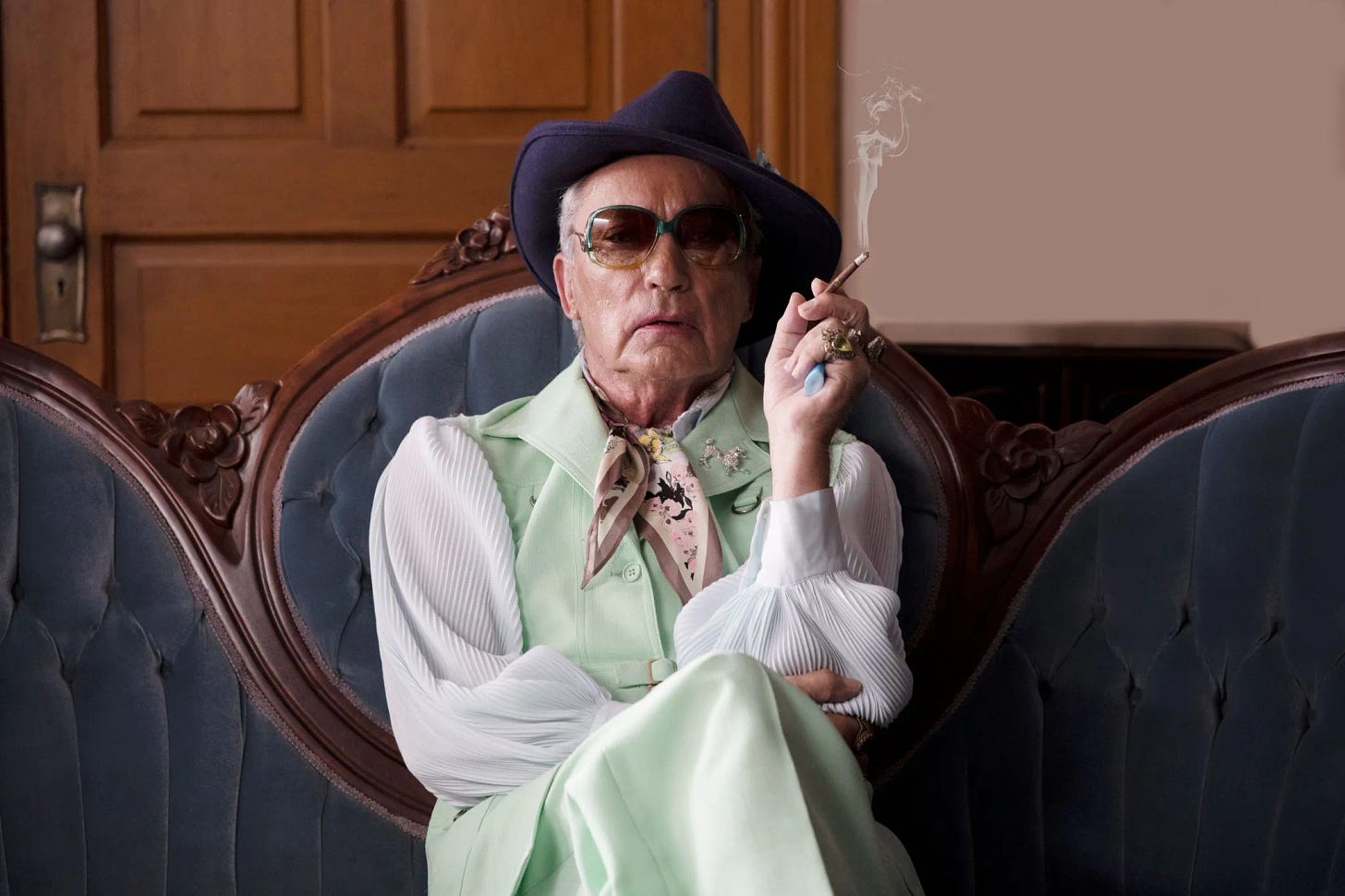 An absolutely fabulously dressed older gay man is sitting in the middle of a grey velvet victorian-style couch. He is wearing a purple fedora, big round tinted glasses, a flowery scarf, a mint-green polyester suit with a white blousey-sleeved shirt underneath. He has giant jeweled rings on each finger of his hand he holds up with a lit and smoking long brown More cigarette. He is facing the viewer with is lipstick-dabbed lips parted...